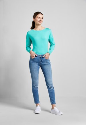 Frau steht mit den Händen in den Taschen, trägt einen türkisfarbenen Pullover, blaue Jeans und weiße Sneakers vor einem schlichten grauen Hintergrund.