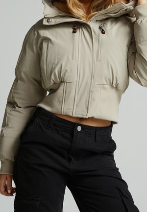 Femme portant une doudoune courte beige avec deux zips sur la poitrine et un pantalon cargo noir taille haute, posant avec une main touchant ses cheveux.