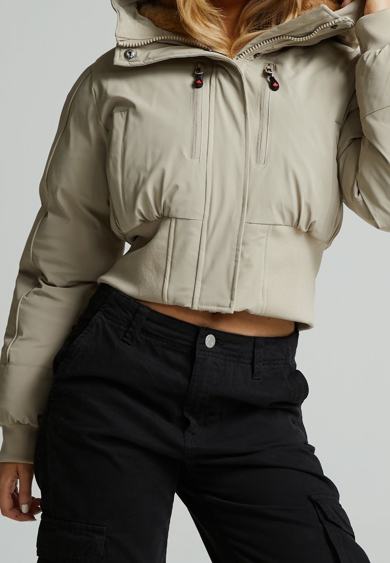 Femme portant une doudoune courte beige avec deux zips sur la poitrine et un pantalon cargo noir taille haute, posant avec une main touchant ses cheveux.