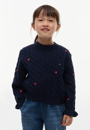 Unge barn med mørkt hår i en hestehale, iklædt en mørkeblå kabelstrikket sweater med små røde broderede mønstre og blå jeans, smilende.