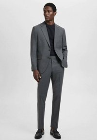 Costum gri croit dintr-un material texturat; design slim-fit, sacou cu un singur nasture, asociat cu o cămașă neagră și pantofi negri.