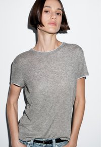 T-shirt grigia a maniche corte con colletto rotondo, caratterizzata da una sottile texture leggera e bordi a contrasto. Indossata con jeans blu.