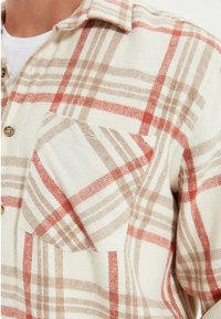 Camisa de cuadros con base crema, con líneas rojas y marrones. Material de franela, cierre de botones y un bolsillo en el pecho.