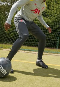 Osoba oblečená ve výcvikovém oblečení Liverpool FC ovládá černý fotbalový míč Nike na travnatém hřišti poblíž brankové sítě.