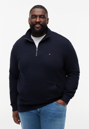 Skægget mand iført en navyblå trøje med kvart-lynlås og lyseblå jeans, stående med den ene hånd i lommen mod en hvid baggrund.