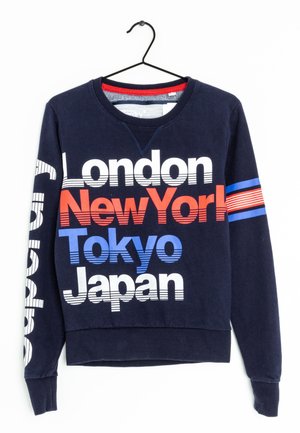 Pull en molleton marine avec un col ras du cou, comportant un texte en gras en blanc, rouge et bleu : "Londres," "New York," "Tokyo," et "Japon." Rayures horizontales sur la manche.