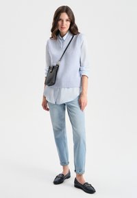 Gilet azzurro lavorato a maglia su una camicia azzurro chiaro con bottoni, abbinato a jeans in denim chiaro e mocassini neri, completato da una piccola borsa nera.