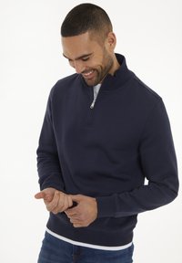 Marineblaue Zip-Up-Sweatjacke mit hohem Kragen, gerippten Bündchen und Saum; über einem weißen T-Shirt getragen; glatte Textur. Hände berühren das Handgelenk.