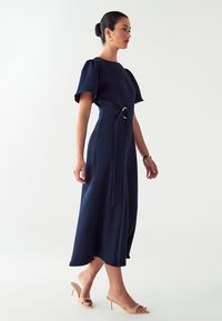 Námornícky modré midi šaty s krátkymi puff rukávmi, opásaný pás s kovovou prackou, vzdušná látka a jemný lesk. Model má na sebe nude topánky.