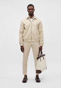 Μπεζ ελαφρύ μπουφάν bomber, μπεζ παντελόνι, σκούρες γόβες και μια κρέμα τσάντα tote με καφέ λεπτομέρειες, σε ένα μοντέλο μπροστά από μια απλή φόντο.