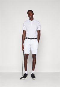 Biely golfový polokošeľa s golierom, v kombinácii s bielymi šortkami a čiernym opaskom. Čierne tenisky a golfová palica v ruke dopĺňajú outfit.