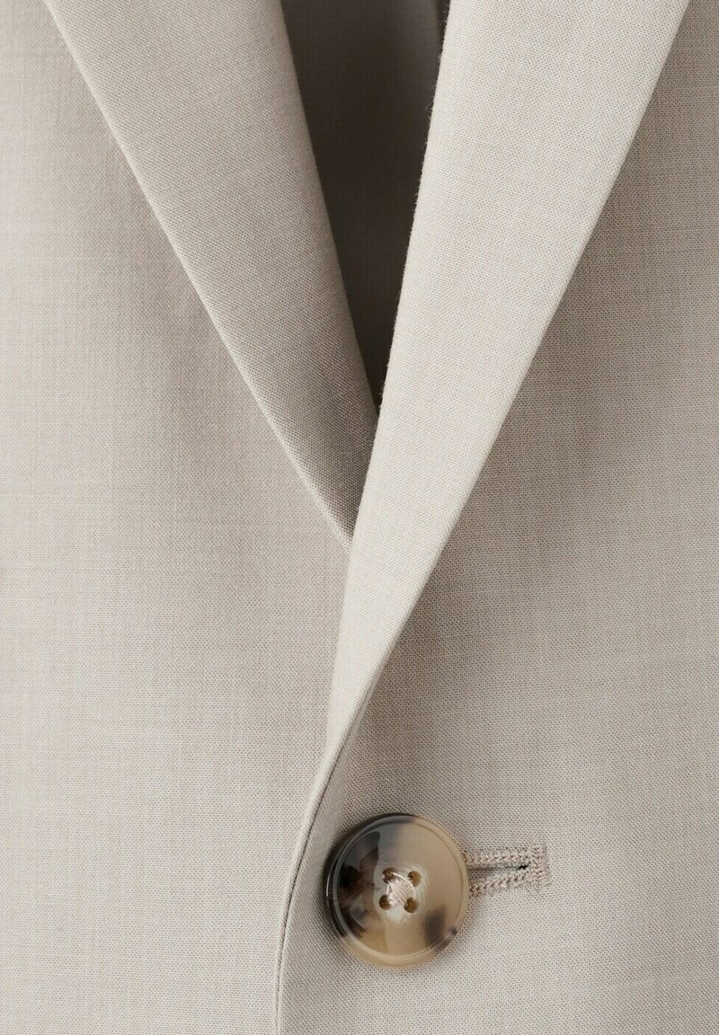 Solapa y cuello de blazer beige con un único botón de carey abrochado en la parte frontal de la prenda.