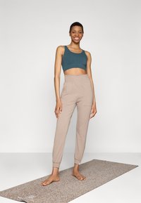 Pantaloni beige con vita elastica e un crop top teal, in piedi su un tappetino da yoga a fantasia con una texture liscia.