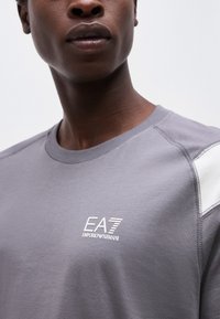 Κοντινό πλάνο ενός ατόμου που φοράει γκρι μπλουζάκι με λευκό λογότυπο "EA7 Emporio Armani" στο στήθος και λευκή ταινία στον ώμο.