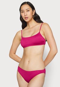 Fuksijos spalvos bikinių komplektas, kuriame yra tekstūruotas braletė su plonomis petnešėlėmis ir atitinkami žemai sėdintys bikini apatiniai. Lengva ir elastinga medžiaga.