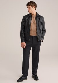 Giacca di pelle nera con chiusura a zip, t-shirt beige sotto, abbinata a pantaloni neri e mocassini neri. Design semplice e su misura.