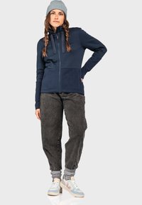 Marineblauer Fleece-Zip-Kapuzenjacke mit strukturierten Einsätzen, graue Hose und helle Sneakers. Modell trägt eine graue Mütze und mehrlagige Socken.