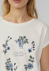 T-shirt en coton blanc avec un motif floral en bleu et vert, comportant une citation en texte centré : "Vous épanouirez si vous prenez le temps de vous arroser."