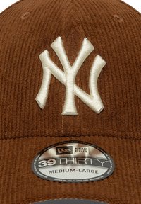 Gorra de pana marrón con un logo bordado en blanco de NY, con una etiqueta negra y plateada que indica tamaño mediano-grande.