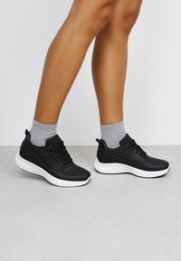 Schwarze Sportschuhe mit glatter Oberfläche, ausgestattet mit einer weißen Sohle, Schnürdesign und Mesh-Details. Getragen mit grauen Socken.