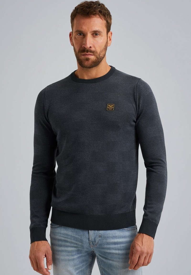 Tmavošedý pletený sveter s rebrovaným golierom a manžetami, na hrudi s malou zlatou emblémou. Úzky strih s hladkou textúrou.