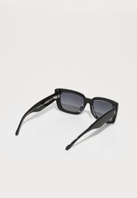 Óculos de sol retangulares pretos com lentes graduadas. Armações grossas e hastes finas, com um pequeno detalhe em metal perto da dobradiça. Textura suave.