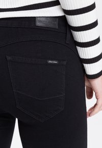 Czarne dżinsy mają tylną kieszeń z poziomym szwem oraz metkę z napisem "Cross Jeans". Tekstura wydaje się gładka i jednolita.