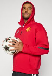 Crvena dukserica na mrežanje sa zatvaračem, s logotipom Manchester Uniteda, crnim akcentima na rukavima i teksturiranom završnom obradom, drži se s bijelom nogometnom loptom.