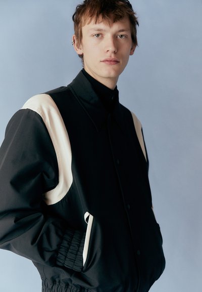 Black jacket with cream accents, snap buttons, and side pockets; features a smooth texture and elastic cuffs, worn over a black turtleneck.