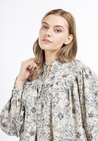Bluse mit Blumenmuster in Creme, mit hohem Ausschnitt, Raffung und weit geschnittenen Ärmeln. Mit Kordelzügen am Kragen.