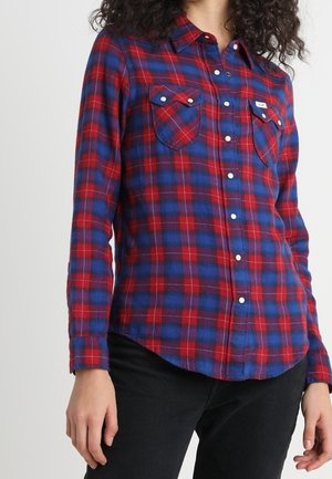 Femme portant une chemise à carreaux rouge et bleue à manches longues avec deux poches poitrine, associée à un pantalon noir.