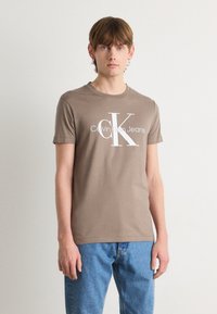 Ruda medvilnė marškinėliai trumpomis rankovėmis, su dideliu baltu "Calvin Klein Jeans" logotipu ir išraiškingu "CK" raidžių dizainu.