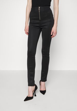 Schmal geschnittene schwarze Hose mit Reißverschluss vorne, kombiniert mit schwarzen spitzen High Heels, gezeigt von der Mitte des Oberschenkels bis zu den Füßen.