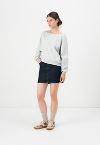 Donna in piedi con le mani in parte nelle tasche, indossa un felpa grigia chiara, una mini gonna in denim scuro, calze beige e scarpe da ginnastica con stampa leopardata.
