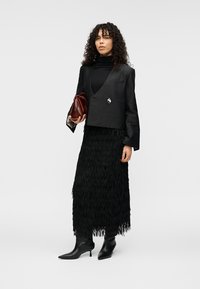 Veste courte noire à finition brillante, associée à une jupe noire à franges. Bottines noires et pochette rouge foncé complètent la tenue.