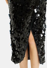 Vestido negro de lentejuelas con una abertura lateral, que presenta lentejuelas circulares dispuestas de manera cercana sobre la tela para un acabado texturizado.