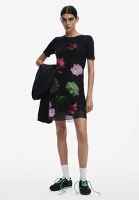 Schwarzes Kurzarmkleid mit buntem Blumenmuster. Das Model hält eine schwarze Jacke und trägt schwarze Sneakers mit grünen Schnürsenkeln und weißen Socken.