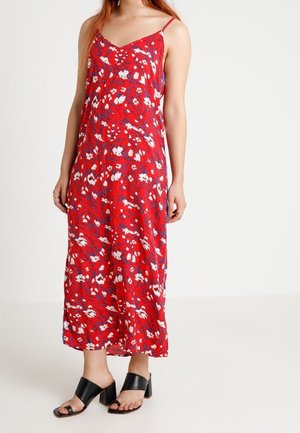 Vrouw die een rode midi-jurk met bloemenprint en dunne bandjes draagt, met zwarte open teen sandalen met hak, staand tegen een effen achtergrond.