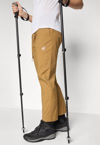 Ανοιχτό καφέ παντελόνια πεζοπορίας με τσέπες, υφή υφάσματος και λογότυπο. Φορεμένα με μαύρες μποτάκες πεζοπορίας και μπατόν πεζοπορίας σε ουδέτερο φόντο.