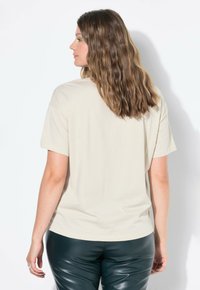 Beżowa bawełniana koszulka o luźnym kroju, z krótkimi rękawami i zaokrąglonym dołem, połączona z czarnymi spodniami przypominającymi skórę, prezentowana z tylnego kąta.