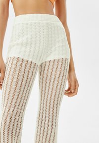 Białe szydełkowe spodnie w wysokim stanie z pionowymi paskami. Materiał ma otwartą strukturę, co daje lekki i przewiewny wygląd.