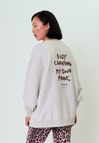 Šedá oversized mikina s kulatým výstřihem, na zádech s hnědým textem: "MÁM ZAPLNĚNO TVORBÍM SVOU VLASTNÍ MAGII." V kombinaci s vzorovanými legínami.