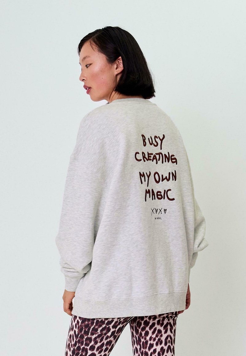 Šedá oversized mikina s kulatým výstřihem, na zádech s hnědým textem: "MÁM ZAPLNĚNO TVORBÍM SVOU VLASTNÍ MAGII." V kombinaci s vzorovanými legínami.