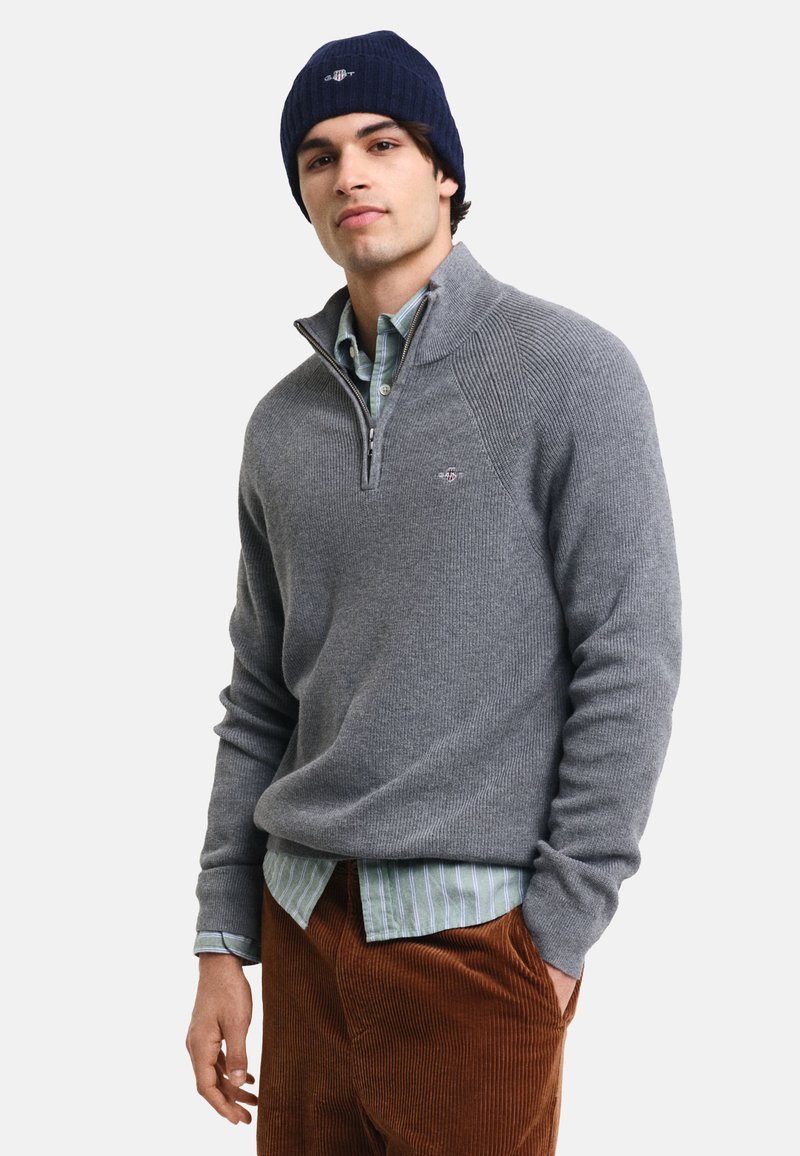 Grauer Strickpullover mit einem Viertel-Reißverschluss-Kragen, ausgestattet mit einer gerippten Struktur. Getragen über einem hellgrünen gestreiften Hemd mit braunen Cordhosen.