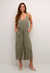 Olíva zöld overall V-kivágással, állítható húzózsinóros derékkal és széles lábszárral. Puha anyagból készült. A modell kezeit a zsebében tartja.