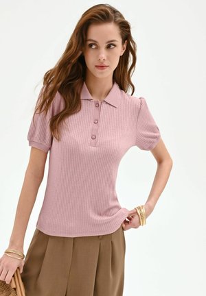 Jeune femme aux longs cheveux portant un polo côtelé rose à manches courtes et un pantalon plissé marron, tenant un sac à main, posant devant un fond blanc.