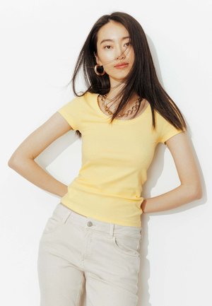 Jeune femme aux longs cheveux foncés portant un haut jaune à manches courtes, un pantalon beige, des boucles d'oreilles créoles dorées et un collier chaîne en or, debout les mains dans le dos.