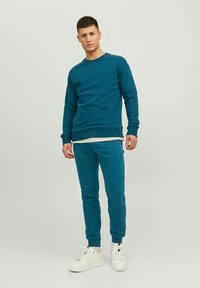 Teal sweatshirt en joggers gemaakt van zachte stof, met ribbels bij de boorden en tailleband, gecombineerd met witte sneakers. Eenvoudig design, geen patronen.