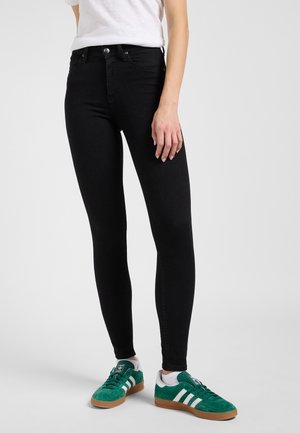 Persoană îmbrăcată în blugi negri mulați, tricou alb și adidași verzi Adidas cu dungi albe, stând pe un fundal simplu, deschis la culoare.