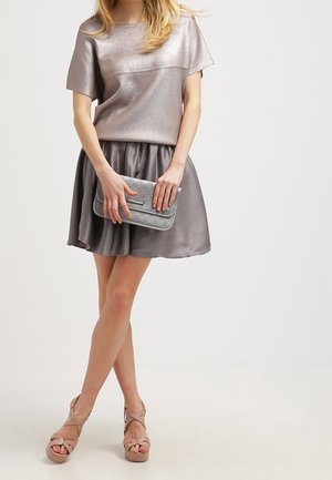 Femme portant un haut métallisé à manches courtes et une jupe assortie, tenant une pochette argentée, chaussée de sandales à plateforme beige avec des brides croisées.